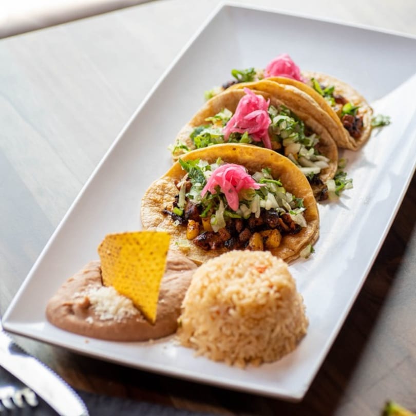 Three Alejandra's Mexican Restaurant tacos with rice and beans on a white rectangular plate.