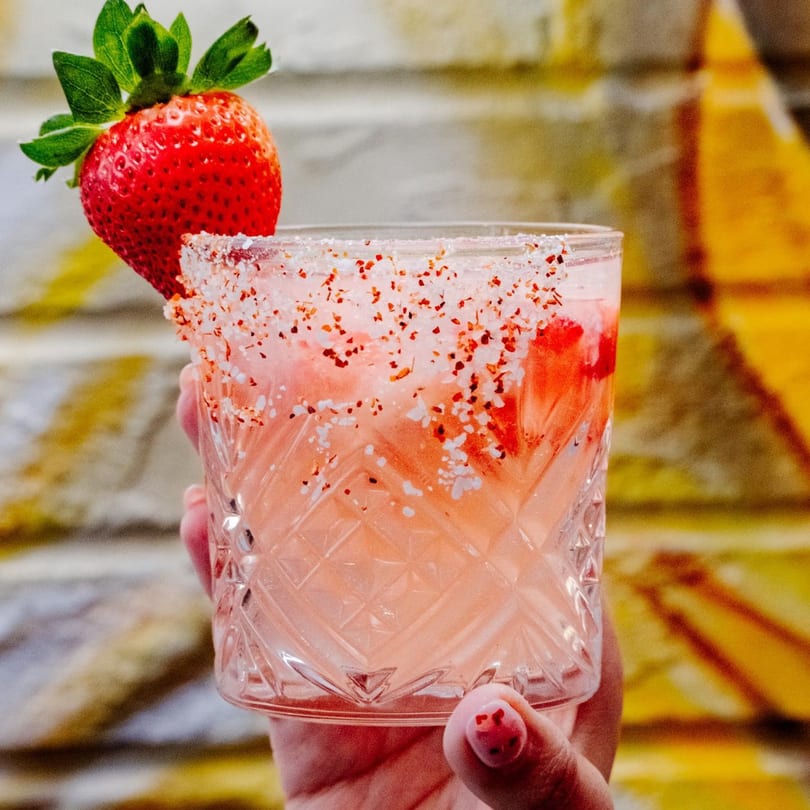 Hand holding a strawberry margarita with a salt and chili rim, garnished with a fresh strawberry.