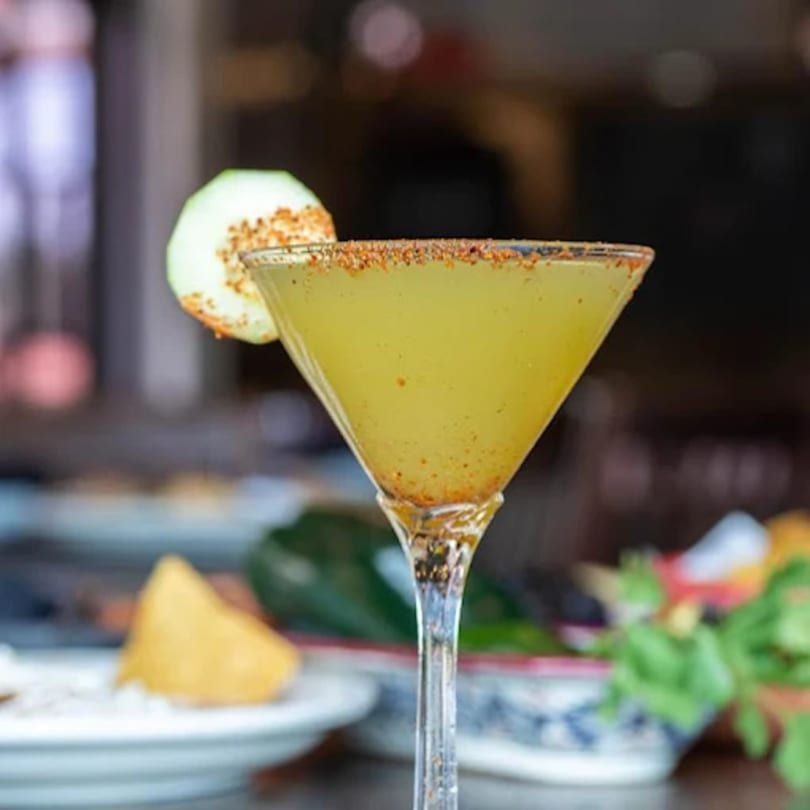 A Mexican margarita in a martini glass, garnished with a cucumber slice and chili powder.