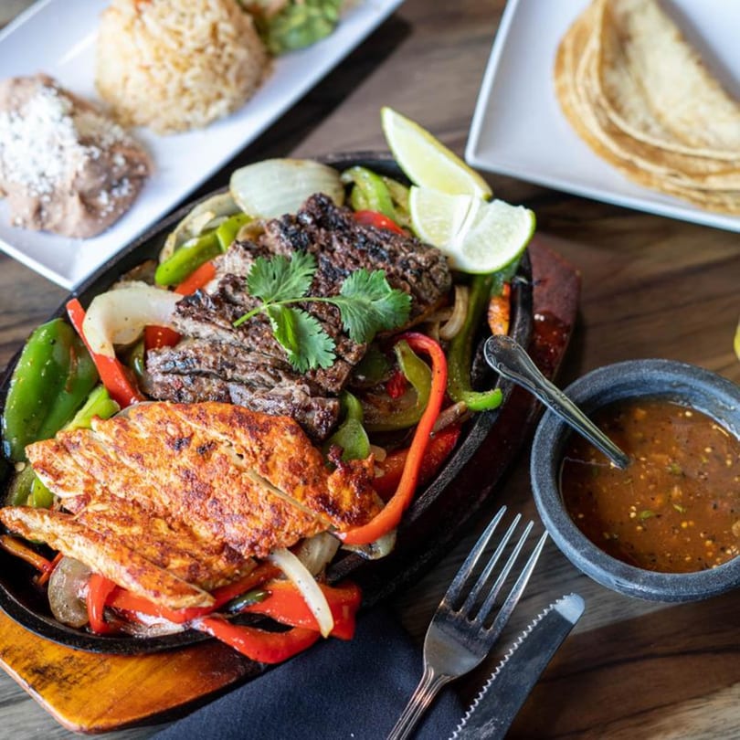 Sizzling fajitas with chicken and steak, bell peppers, onions, limes, and a side of salsa.