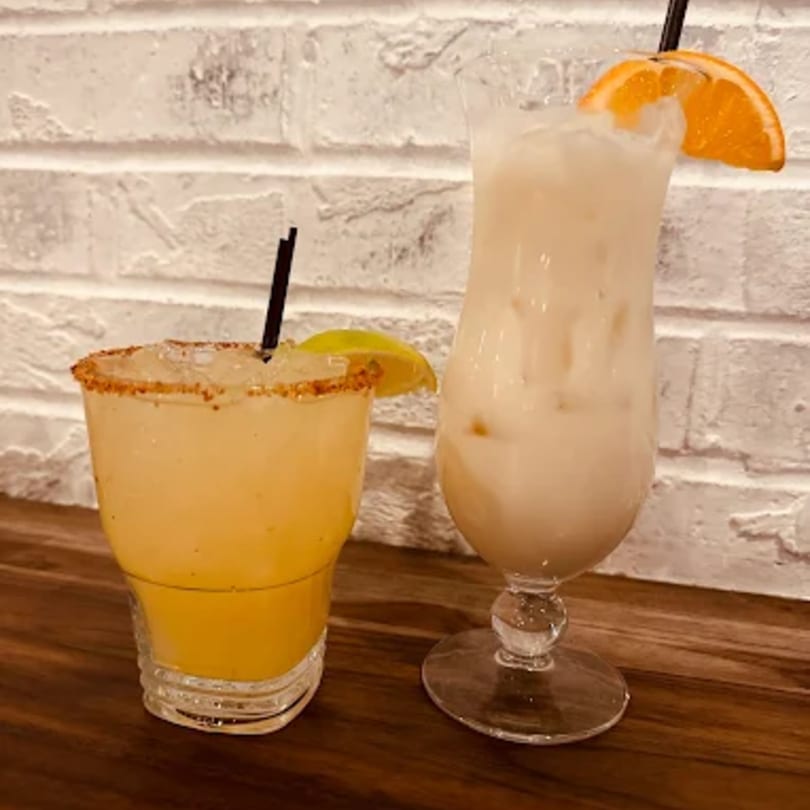 Two drinks, a margarita with a lime and a creamy drink with an orange slice, on a wooden table.
