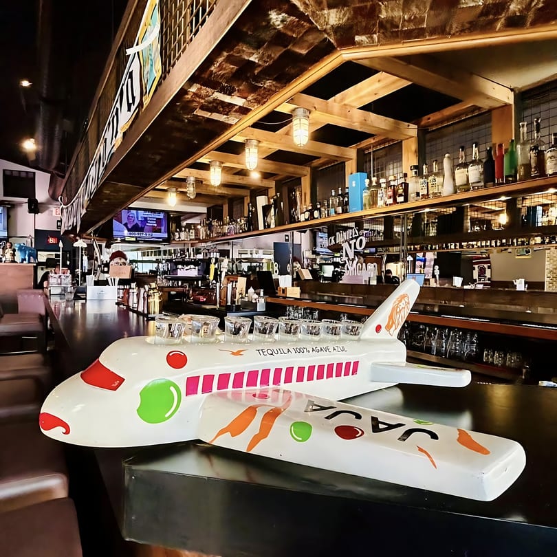 A white airplane-shaped tequila shot holder with colorful accents sits on a dark bar counter.