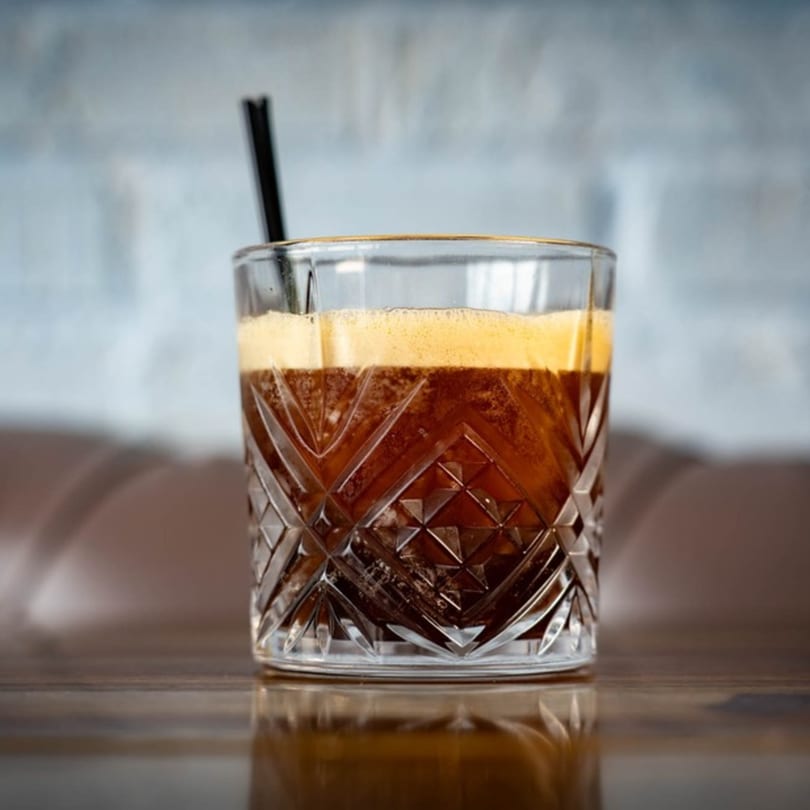 A dark, foamy drink in a cut glass with a black straw on a wooden table.