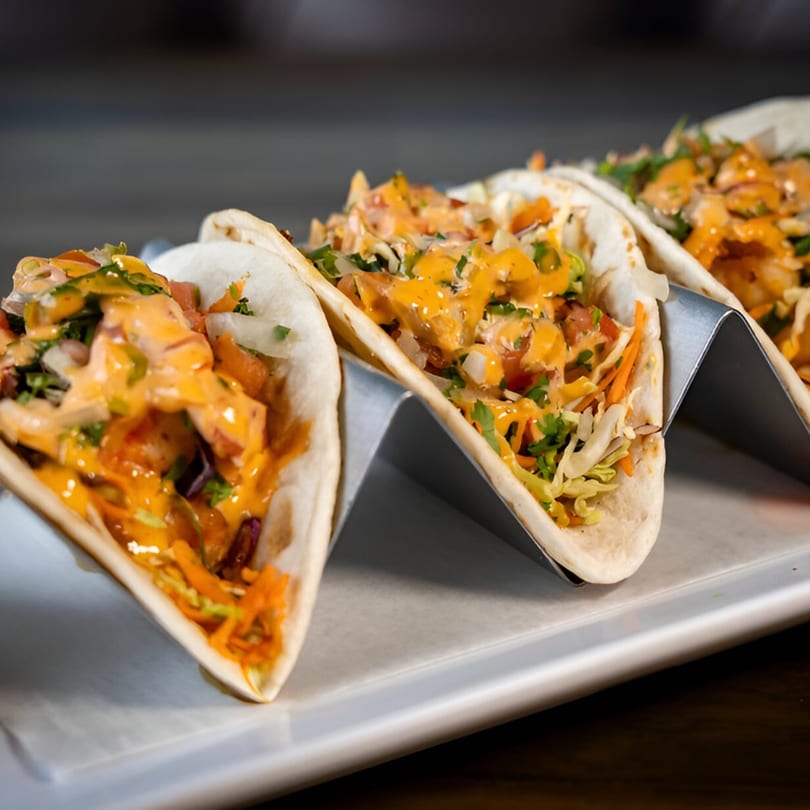 Three Alejandra's Mexican Restaurant tacos with orange sauce, shredded lettuce, and carrots in metal holders.